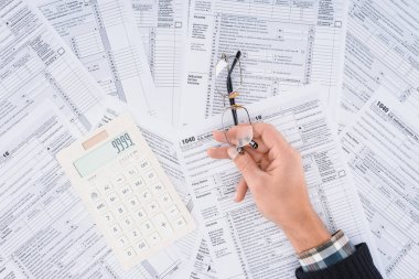 cropped view of man holding glasses with calculator and tax forms on background clipart