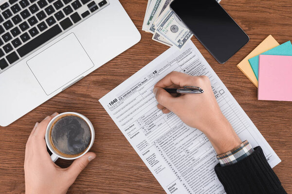 cropped view of man holding coffee cup and filling tax form at workplace with digital devices