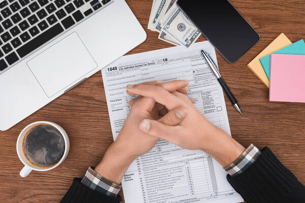 partial view of man with folded hands sitting at desk with tax form and laptop