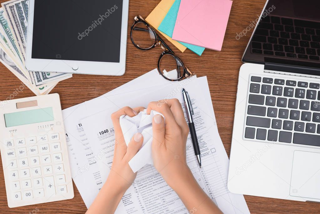 cropped view of woman holding crumpled paper ball at desk with tax forms and digital devices