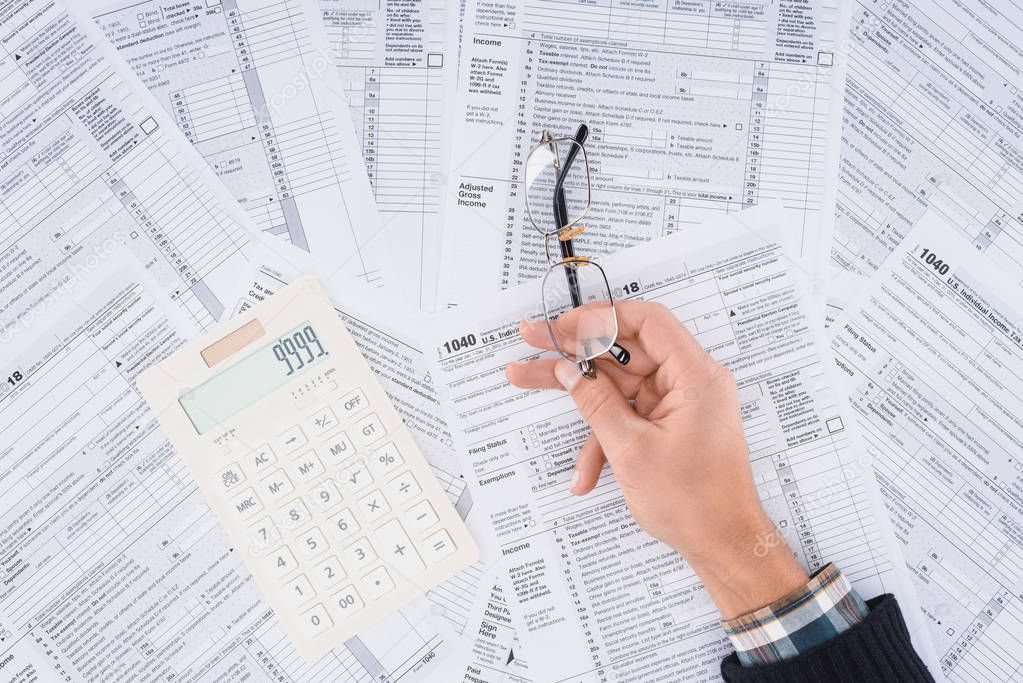 cropped view of man holding glasses with calculator and tax forms on background