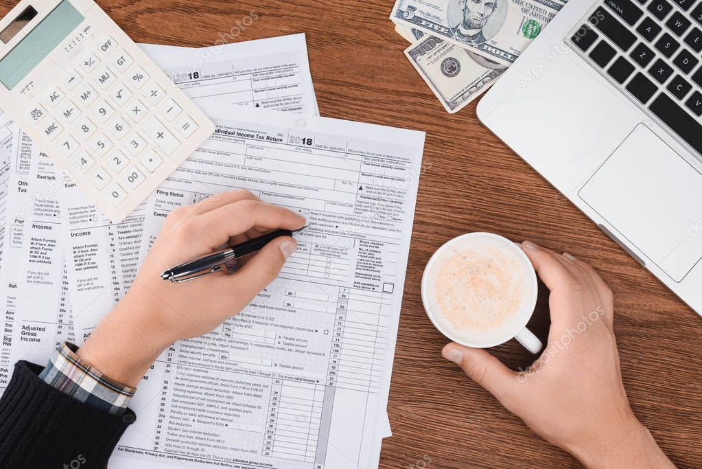 cropped view of man holding coffee cup and filling tax forms at workplace
