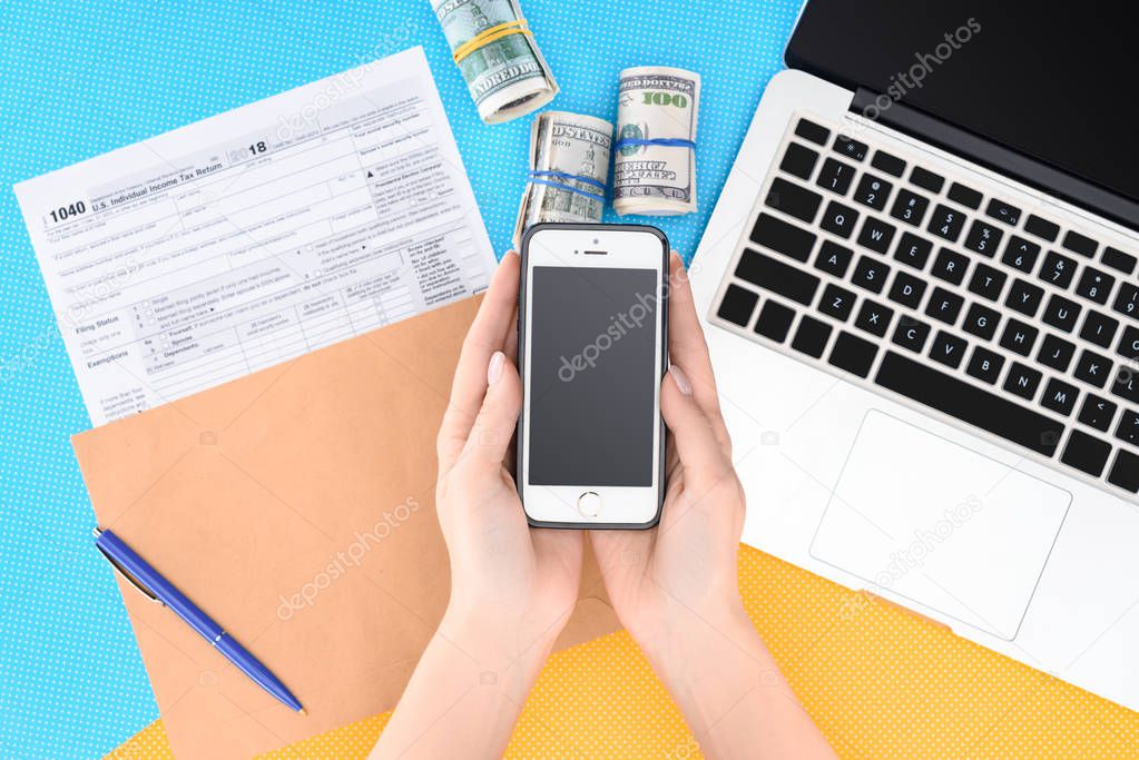 cropped view of woman holding smartphone with blank screen at workplace with laptop, tax form and money rolls on background