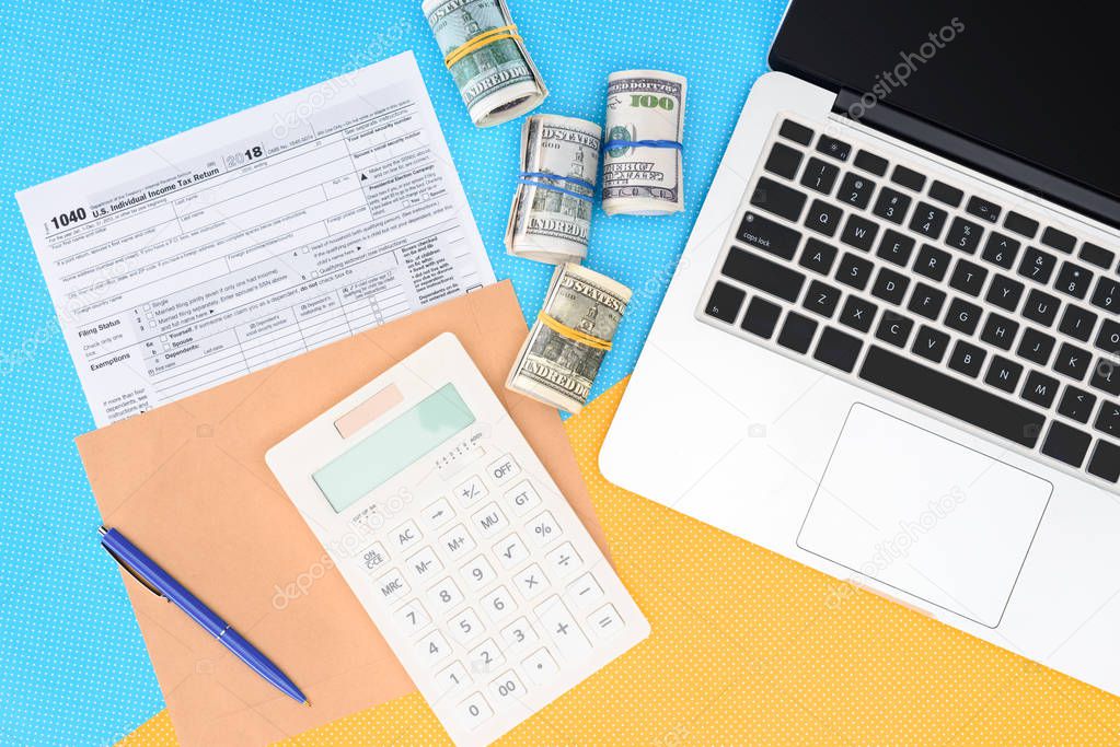 top view of tax form, money rolls and laptop on blue and yellow background