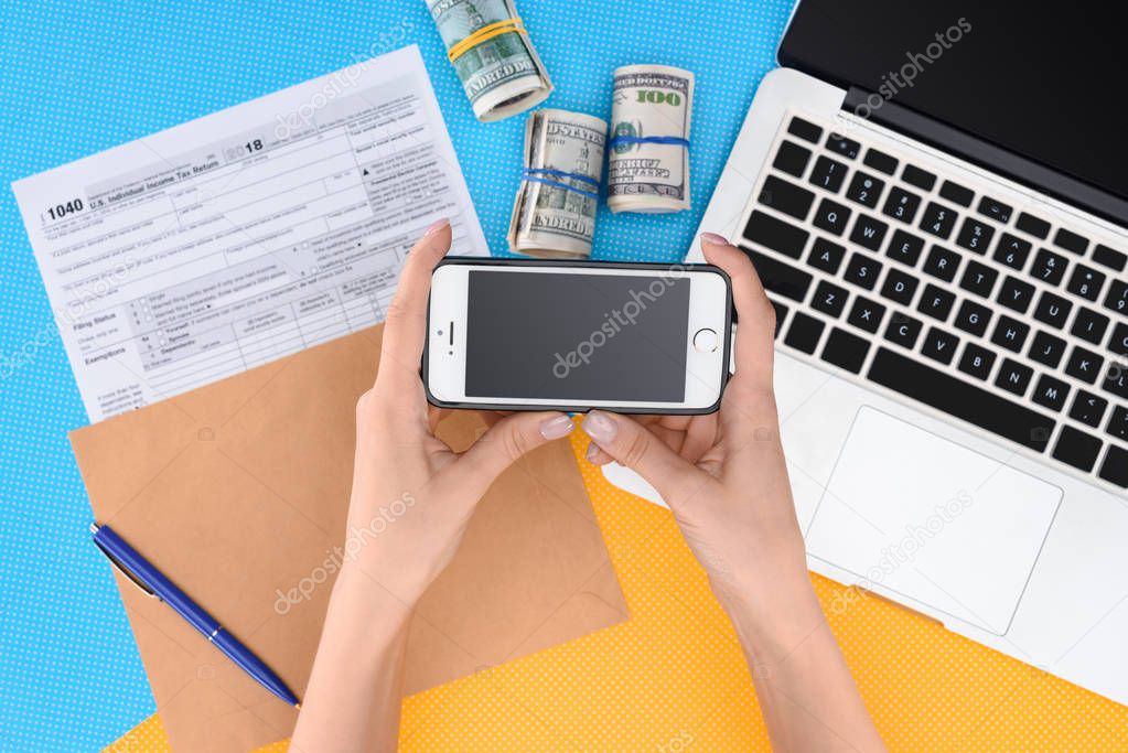 partial view of woman holding smartphone with blank screen at workplace with laptop, tax form and money rolls on background