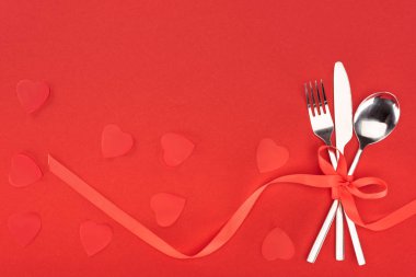 elevated view of cutlery wrapped by festive ribbon near heart symbols isolated on red, st valentine day concept clipart