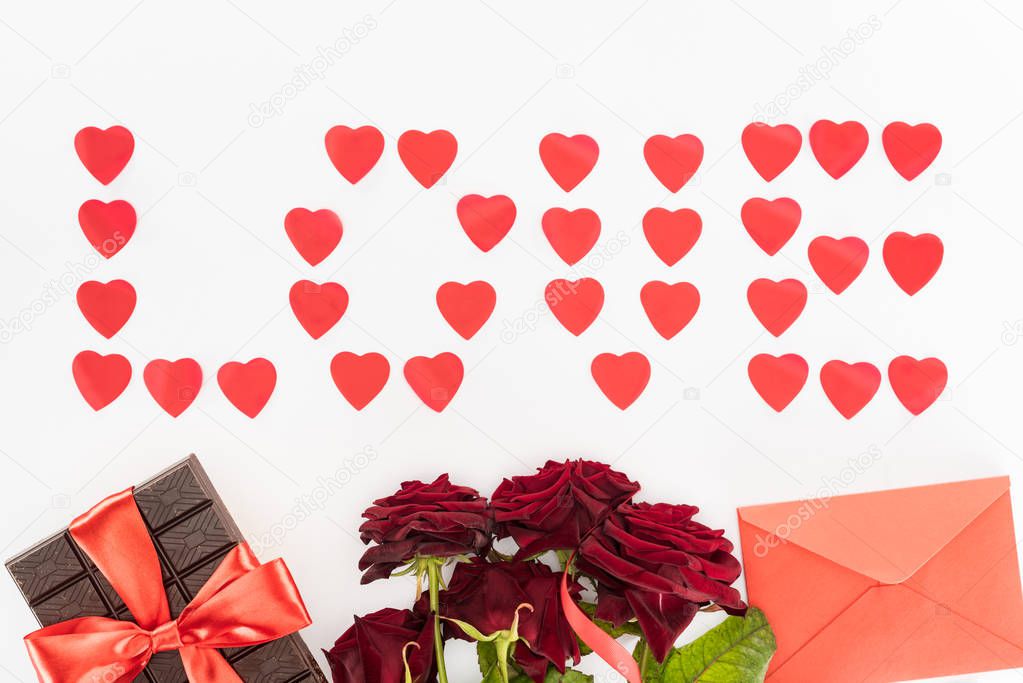 top view of lettering love made of heart symbols, chocolate wrapped by festive ribbon, red roses and envelope isolated on white, st valentine day concept