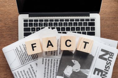 top view of wooden cubes with word fact and newspapers on laptop on wooden tabletop clipart