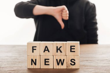 cropped image of journalist showing thumb down to wooden cubes with words fake news at wooden table clipart