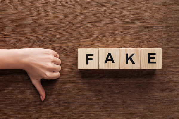 cropped image of journalist showing thumb down to wooden cubes with word fake at wooden table