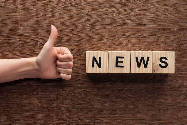 cropped image of journalist showing thumb up to wooden cubes with word news at wooden table