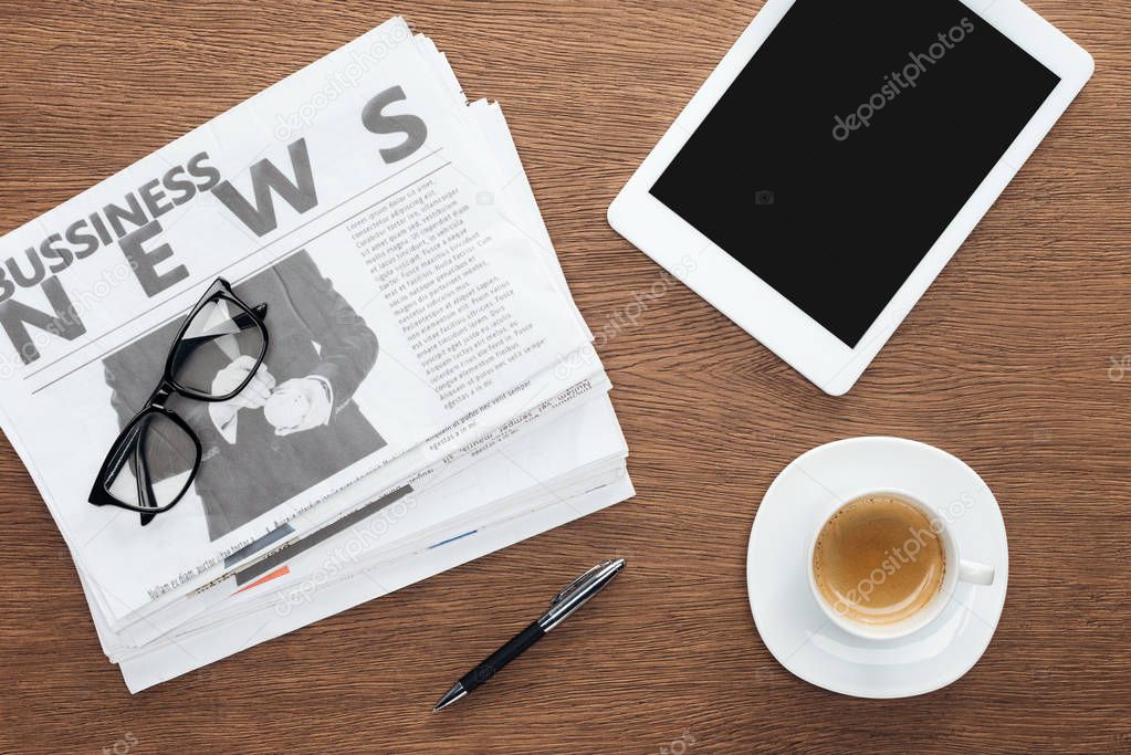 elevated view of cup of coffee, pen, tablet with blank screen and business newspapers on wooden tabletop