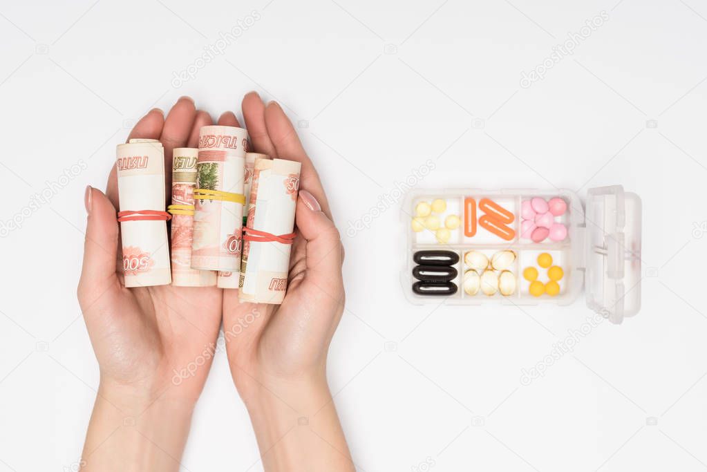 cropped view of woman holding money rolls in hands near plastic container with pills isolated on grey
