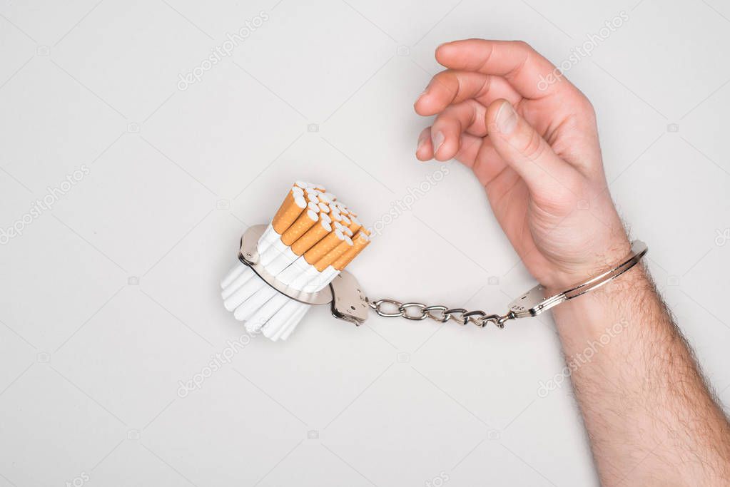 Partial view of man in handcuffs posing with cigarettes isolated on grey, nicotine addiction concept