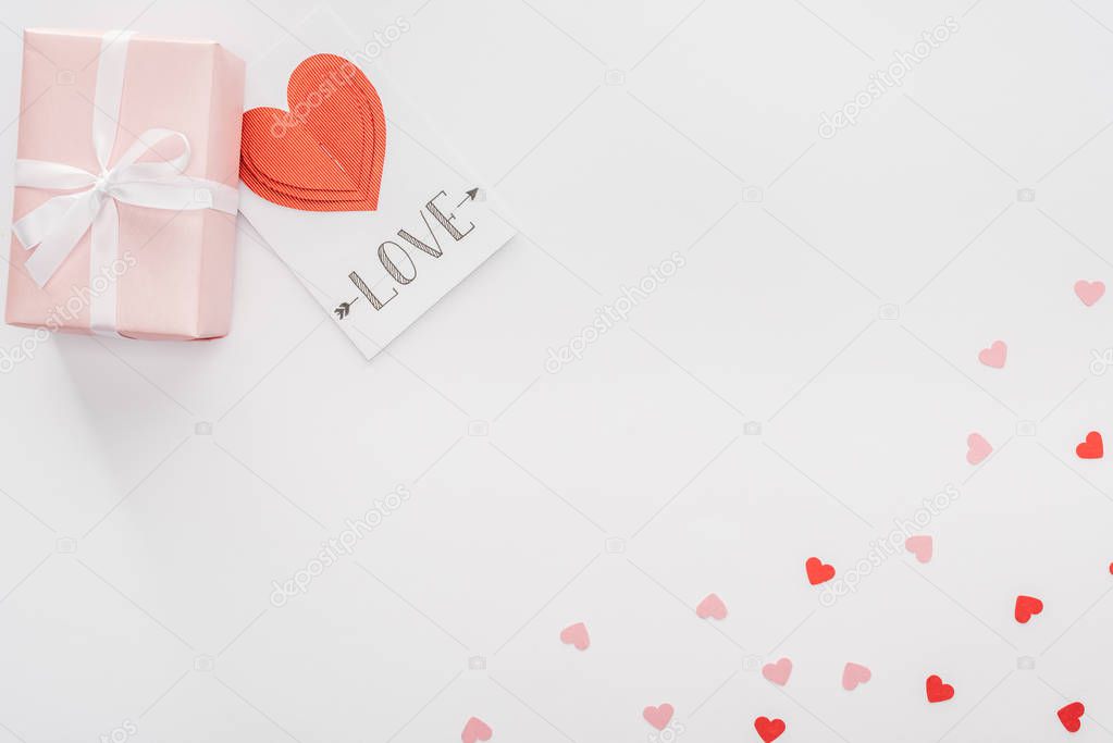 top view of gift box, paper hearts and greeting card with 'love' lettering isolated on white, st valentines day concept