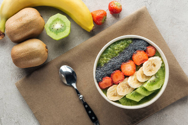 top view of smoothie bowl with fresh fruits, spoon and ingredients on grey background