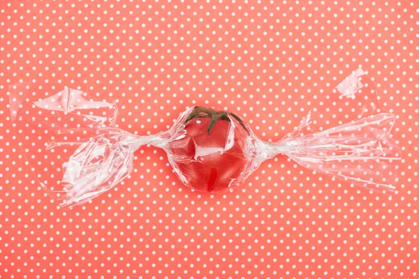 top view of red fresh tomato in transparent candy shaped wrapping on red dotted background