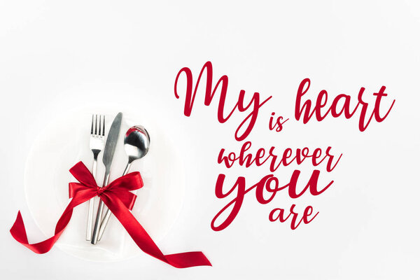 elevated view of fork, knife and spoon wrapped by red festive bow on plate isolated on white, st valentine day concept with "my heart is wherever you are" lettering