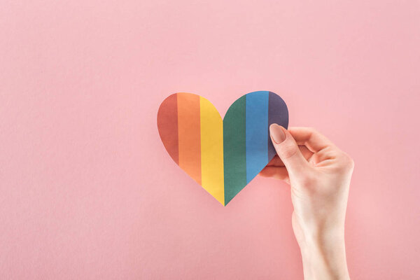 partial view of woman holding rainbow colored paper heart on pink background, lgbt concept