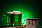 glasses of green irish beer near golden coins and cube calendar on green background