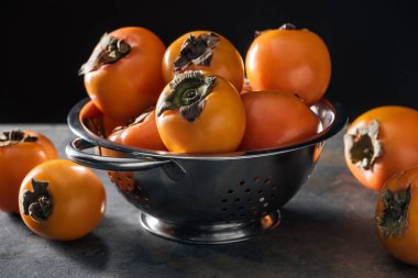 orange and ripe persimmons in colander isolated on black clipart