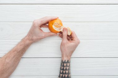 partial view of tattooed man peeling tangerine on white wooden surface clipart