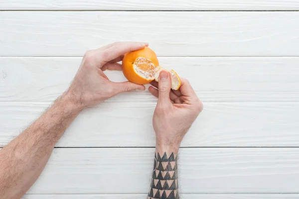 partial view of tattooed man peeling tangerine on white wooden surface