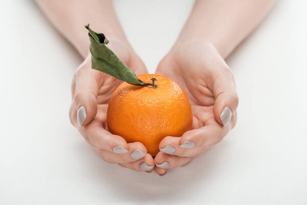 partial view of woman holding ripe whole unpeeled tangerine with leaf on white background