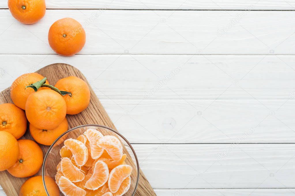 top view of peeled tangerine slices in glass bowl and whole ripe tangerines on wooden chopping board 