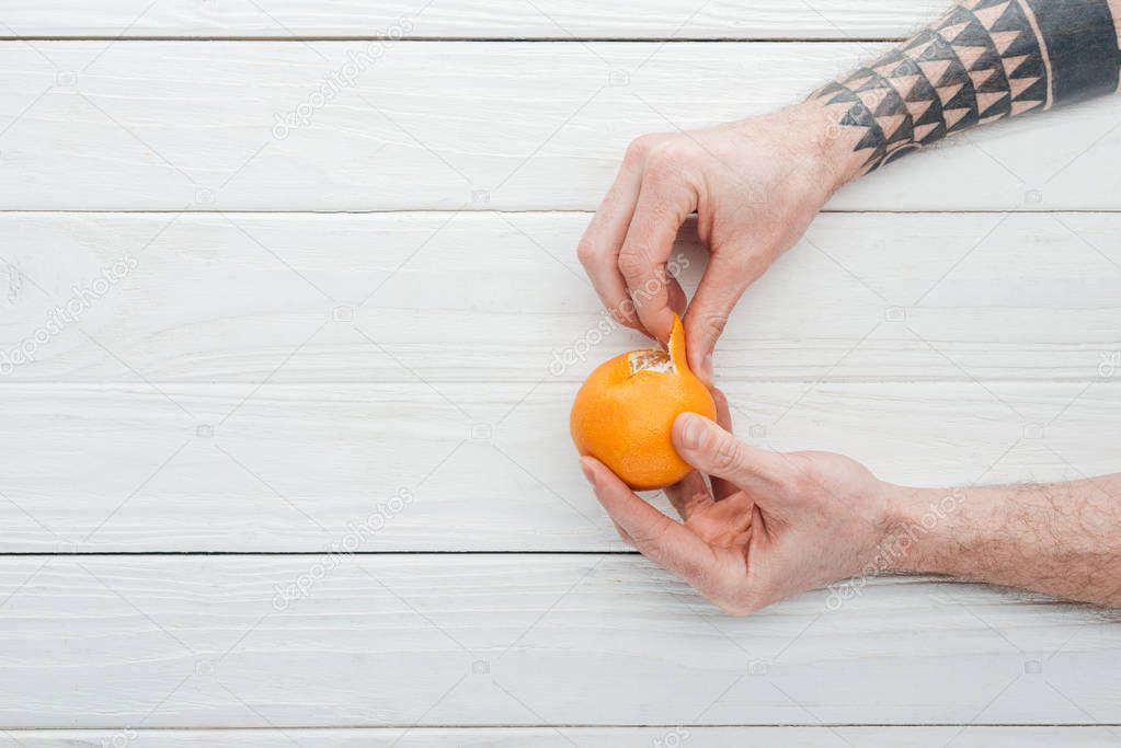 cropped view of tattooed man peeling tangerine on white wooden background