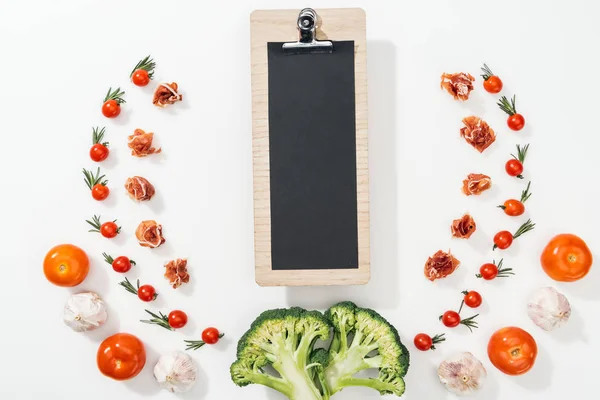 top view of clipboard among tomatoes, leaves, broccoli, prosciutto and garlic