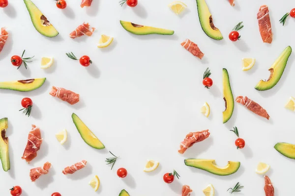 flat lay of prosciutto near sliced avocados, lemons and cherry tomatoes on white background