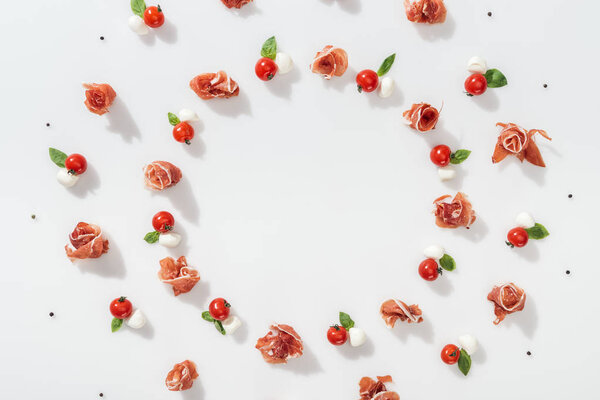 flat lay of tasty prosciutto near red cherry tomatoes, mozzarella cheese and peppercorns on white background