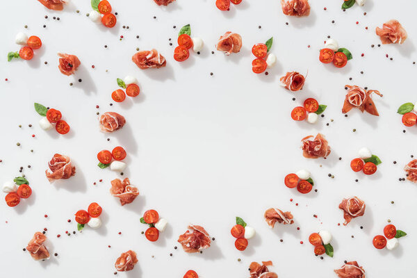 flat lay of prosciutto near red cherry tomatoes, mozzarella cheese, green basil leaves and peppercorns on white background