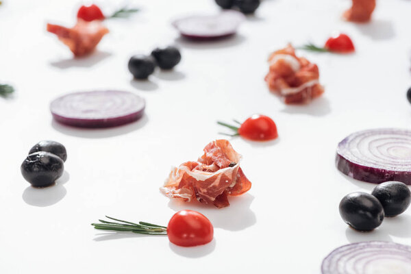 selective focus of prosciutto near cherry tomatoes with rosemary twigs near red onion rings and black olives on white background