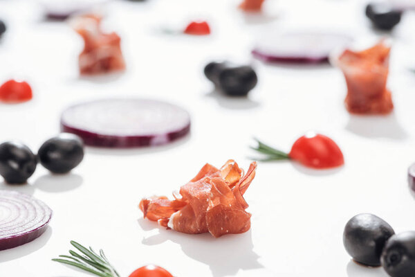 selective focus of delicious prosciutto near cherry tomatoes with rosemary twigs and red onion rings on white background