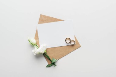 top view of envelope with empty card, flowers and golden wedding rings on grey background clipart