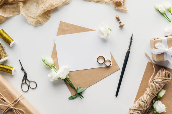 вид сверху на пустую открытку с конвертом, чернилами, эустомой, штампом, завернутыми подарками, ножницами, бечевками и золотыми обручальными кольцами на сером фоне
