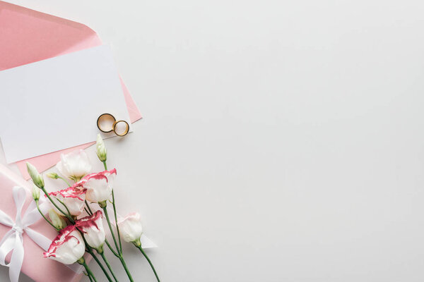 top view of empty card with pink envelope, flowers, wrapped gift and golden wedding rings on grey background