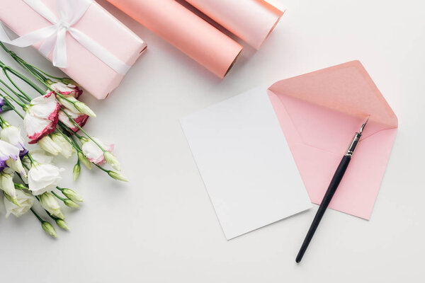верхний вид розового конверта с пустой карточкой и чернилами, цветы, завернутый подарок, рулоны бумаги на сером фоне
