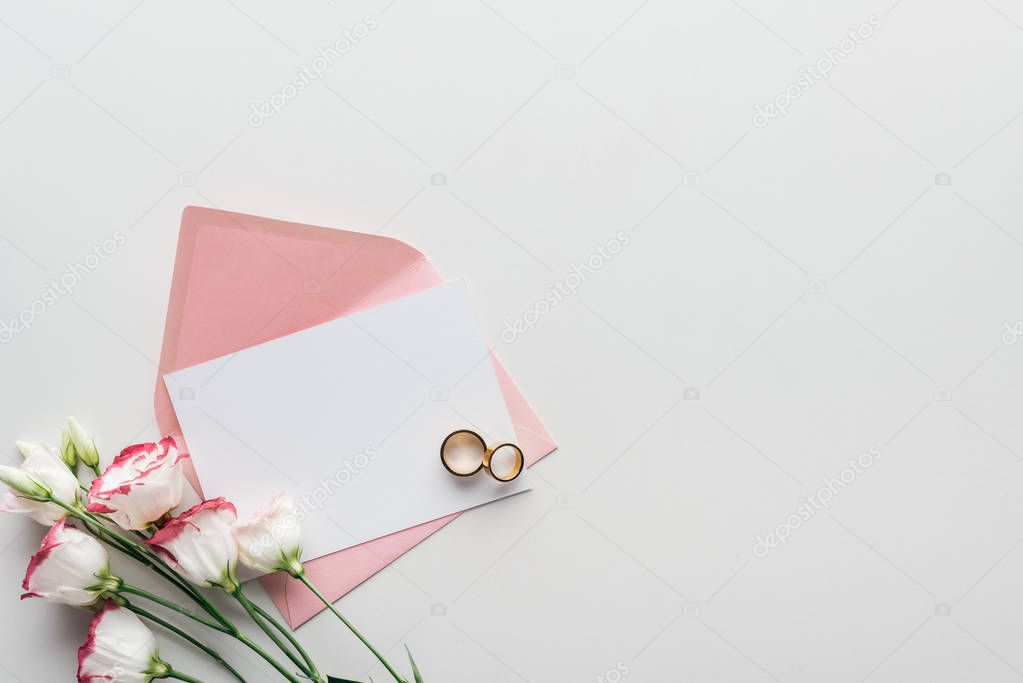 top view of empty card with pink envelope, eustoma and golden wedding rings on grey background