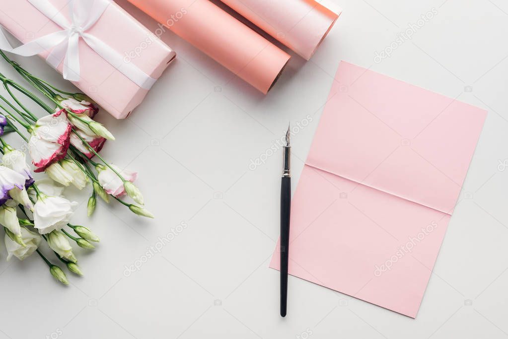 top view of pink empty card with ink pen, flowers and rolls of paper on grey background
