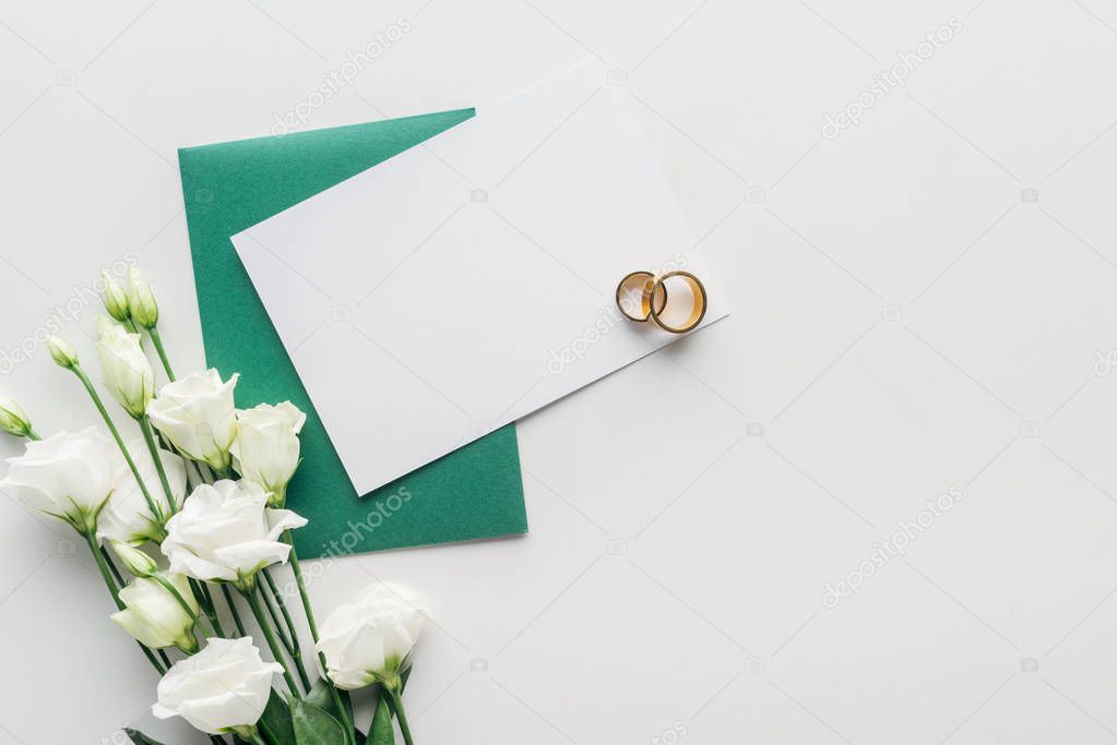 top view of empty card with green envelope, flowers and golden wedding rings on grey background