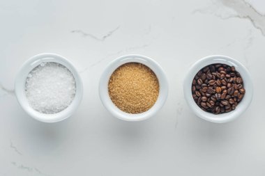 top view of bowls with salt, coffee grains and brown sugar on white surface clipart