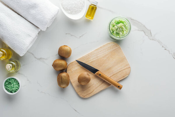 верхний вид стола для резки дерева с ножом и киви, полотенца и различные компоненты для ручной косметики на белой поверхности
