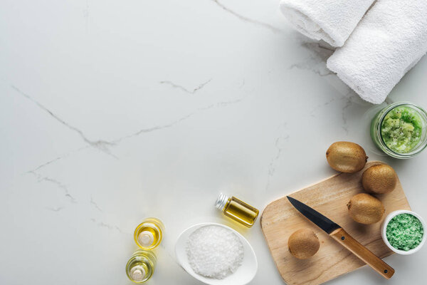 верхний вид миски и контейнеров с косметическими компонентами, полотенца, разделочный стол с ножом и киви на белой поверхности

