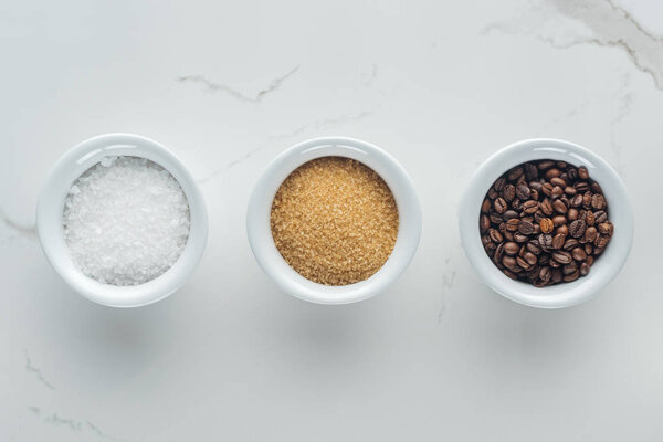 верхний вид миски с солью, зерном кофе и коричневым сахаром на белой поверхности
