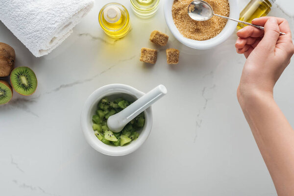 cropped view of woman holding spoon of brown sugar, pounder with kiwi, and various cosmetic ingredients on white surface