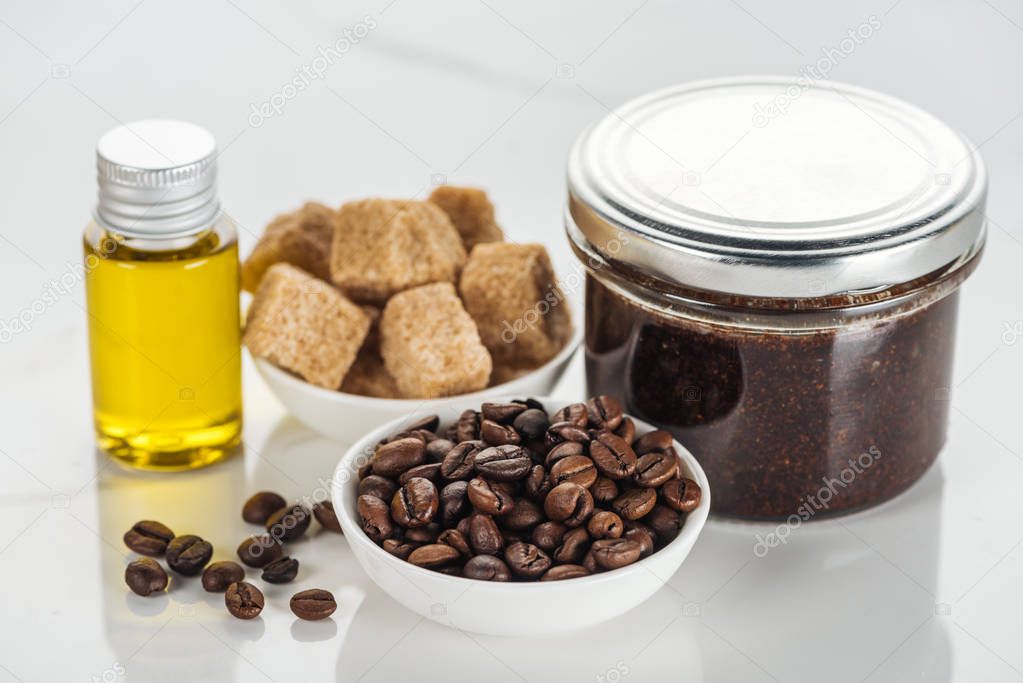 selective focus of bowls with brown sugar cubes and coffee grains, and glass containers with ground coffee scrub and oil on white surface