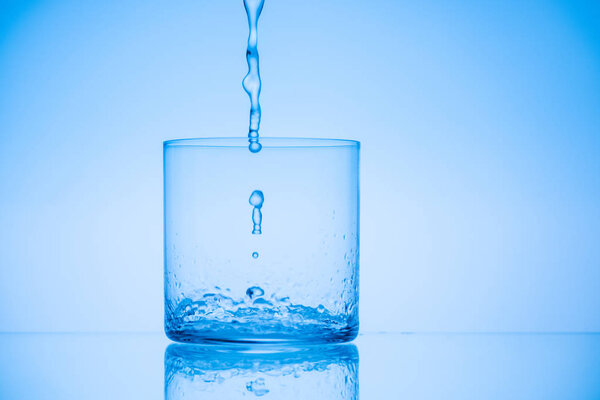 toned image of water pouring in empty glass on blue background
